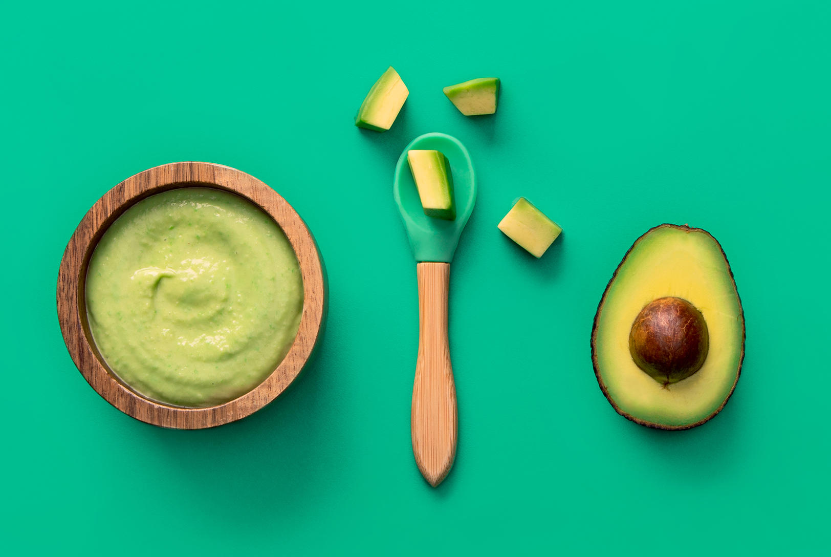 Avocado purée in a small wooden bowl, with a baby spoon, surrounded by avocado chunks and half an avocado on a bright teal background.