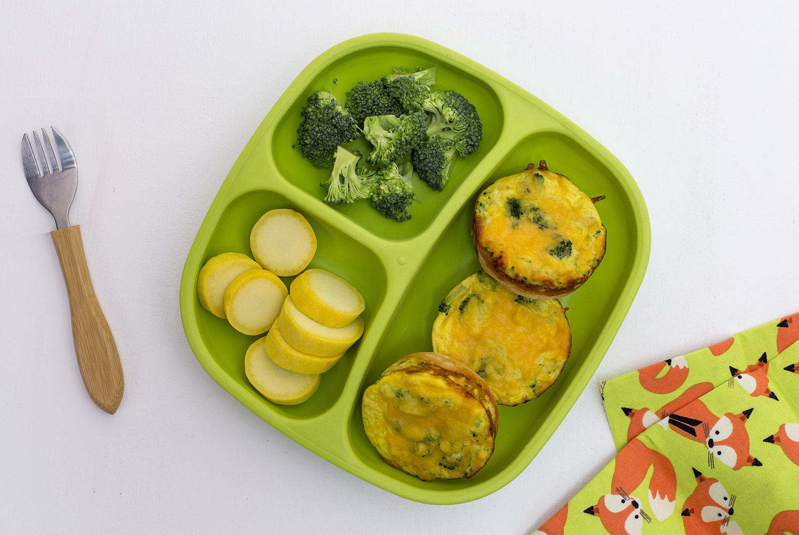 Three Mini Broccoli and Cheese Quiches on a green divided plate, with steamed broccoli and sliced yellow squash, next to a child's fork.
