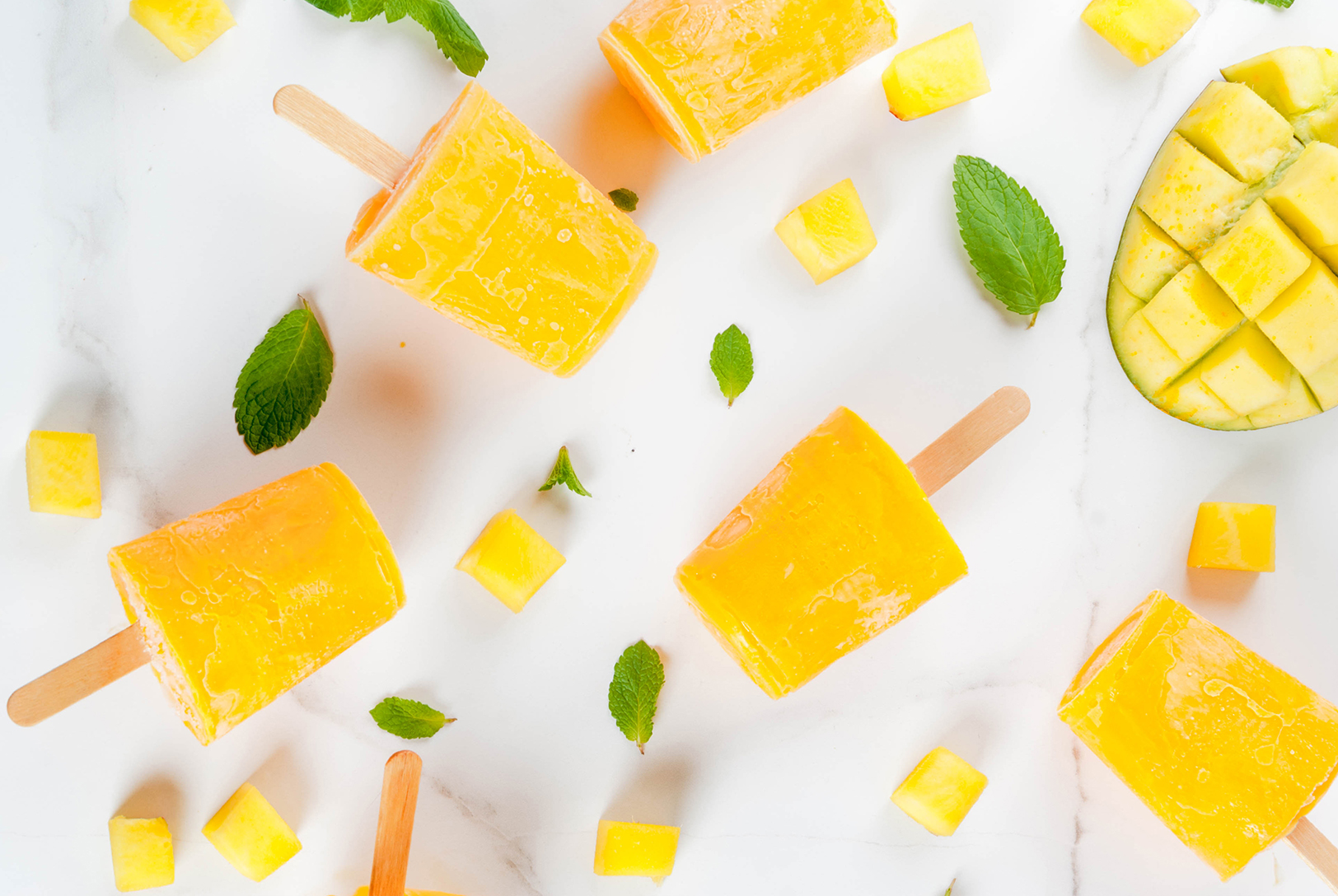 Several bright yellow mango and banana popsicles scattered on a white marble surface, along with chunks of fresh mango and green mint leaves.