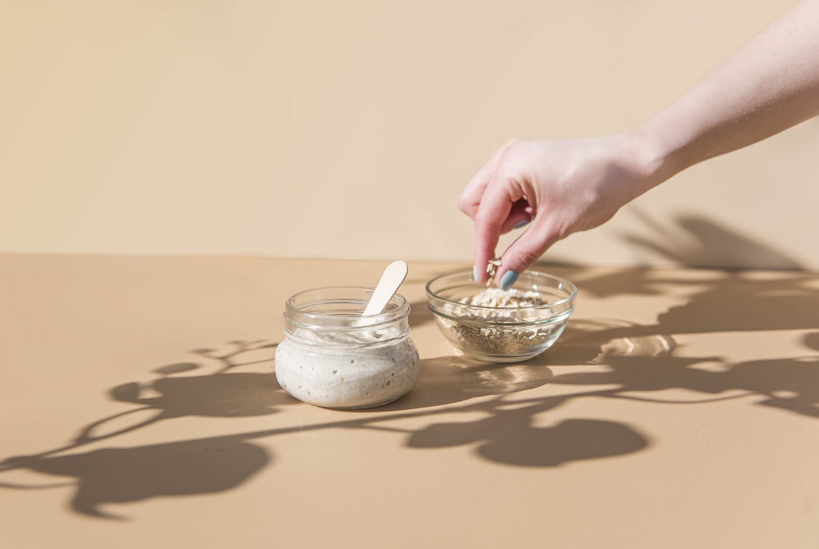 A small glass jar with Moisturizing Oat Face Mask with a wooden spatula, next to a glass bowl of dry oats being picked by a hand.