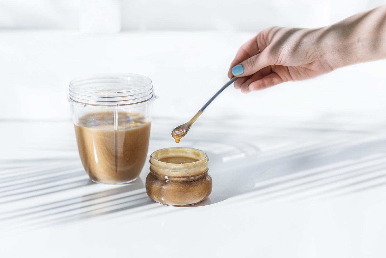 A small glass jar filled with Body Sugar Scrub, with a hand scooping some out with a spoon, next to a blender cup with more scrub.