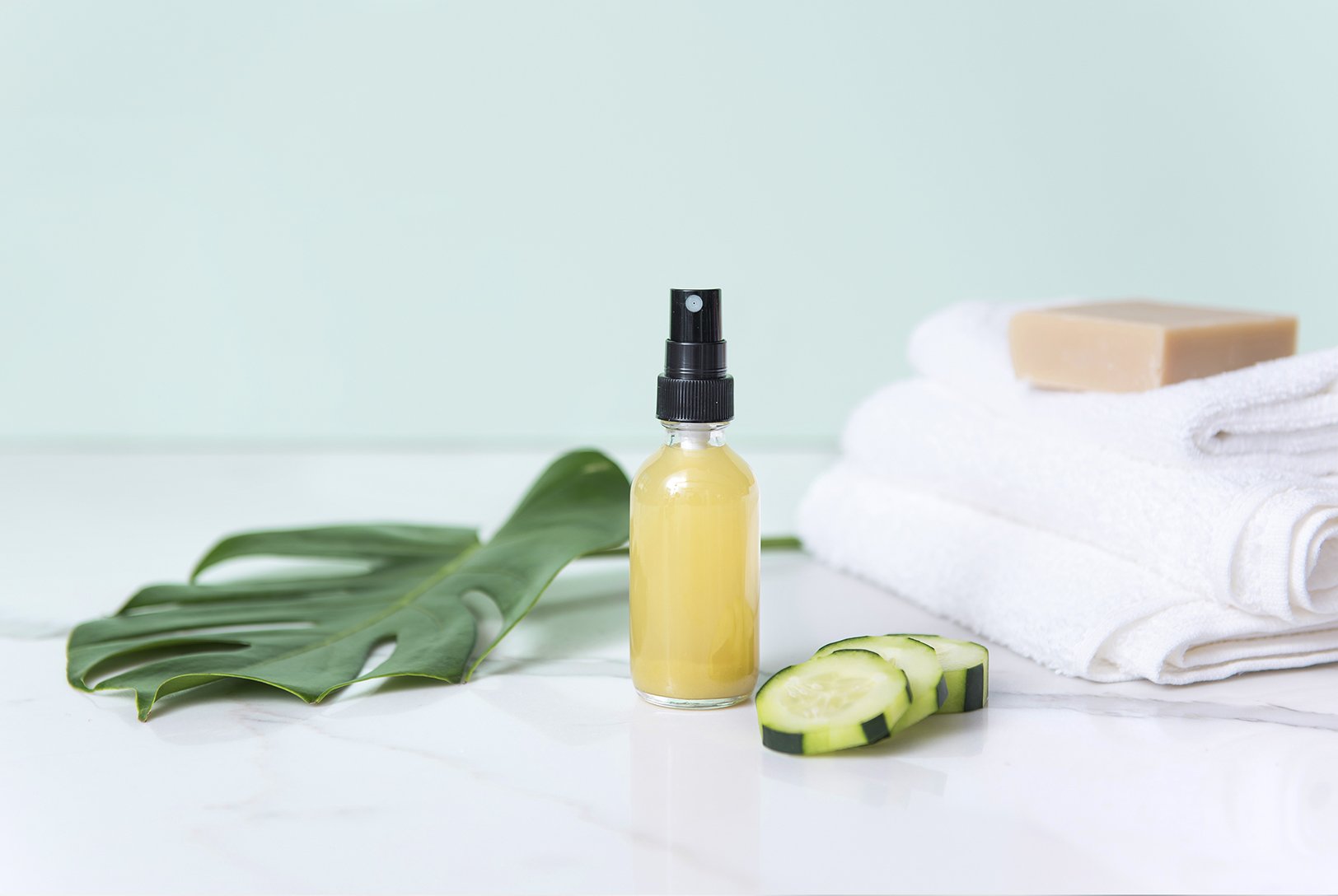 A spray bottle filled with Green Tea and Cucumber Toning Mist, next to slices of cucumber and a green leaf, with a stack of white towels and a bar of soap.