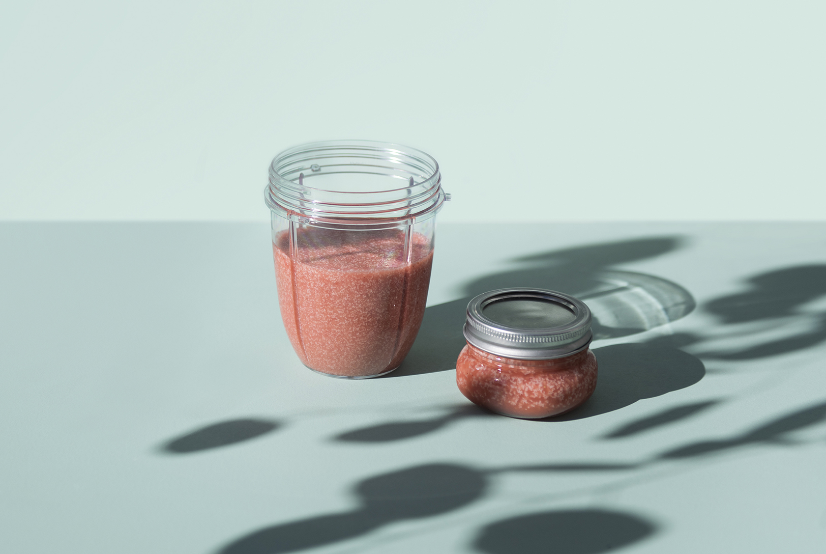 A nutribullet cup partially filled with Strawberry Hair Mask, next to a small glass jar with more of the mask, on a blue-green surface with shadows.