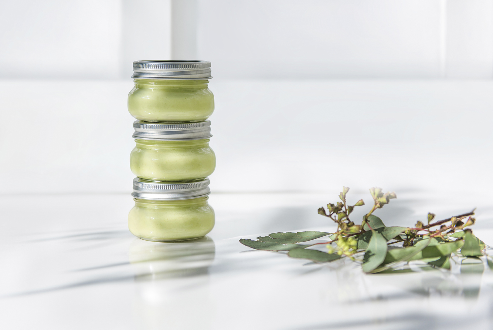 Three small stacked glass jars containing Avocado Banana Mask, next to a sprig of leaves on a white marble surface.