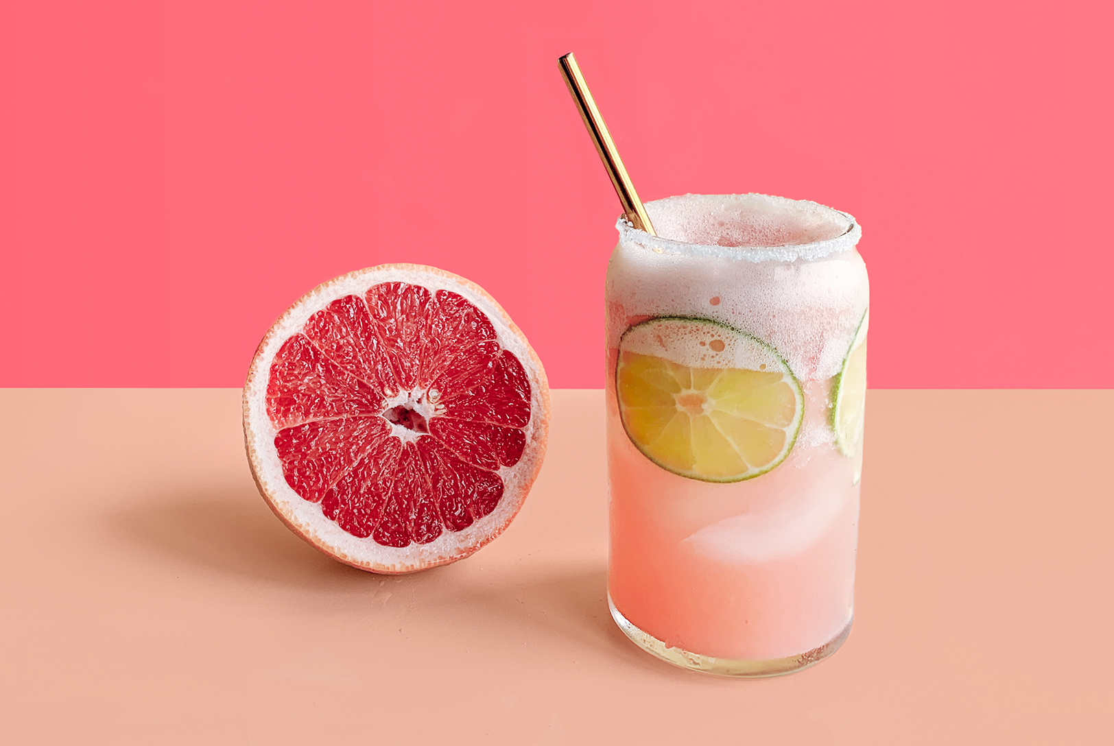 Paloma drink in a glass with a gold metal straw with citrus slices next to a halved grapefruit.