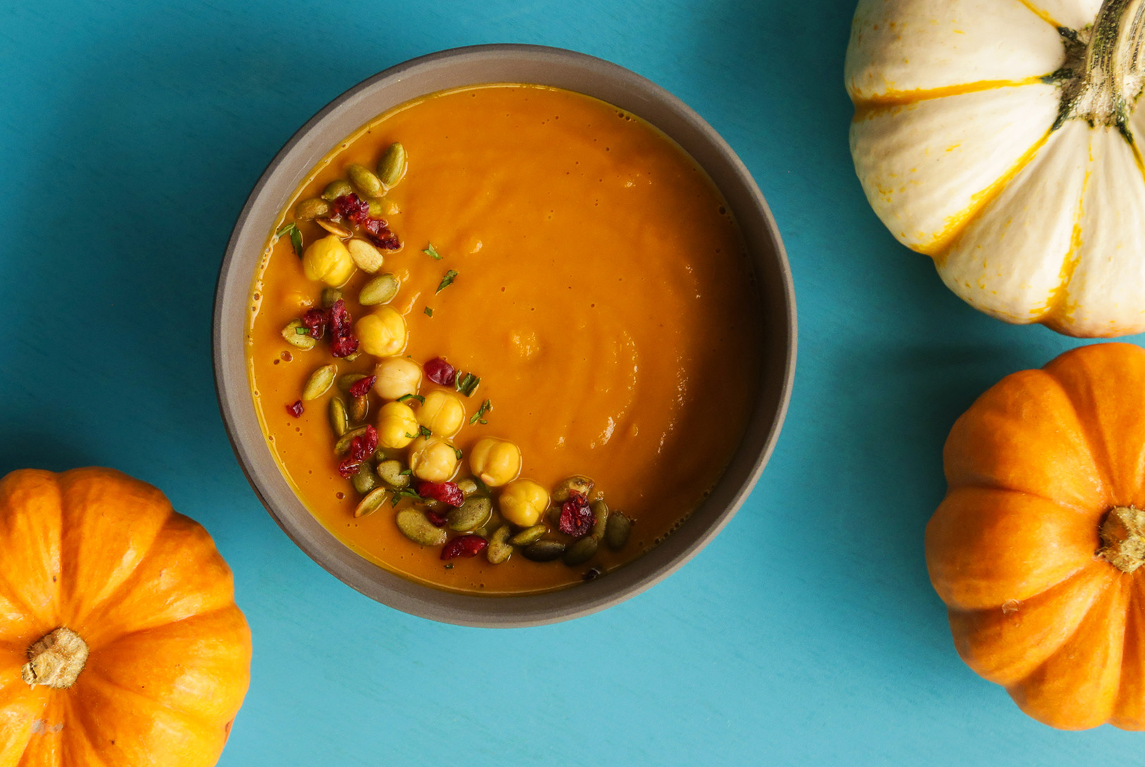 Pumpkin soup served in a bowl garnished with chickpeas, seeds and pomegranate surrounded by whole pumpkins. 