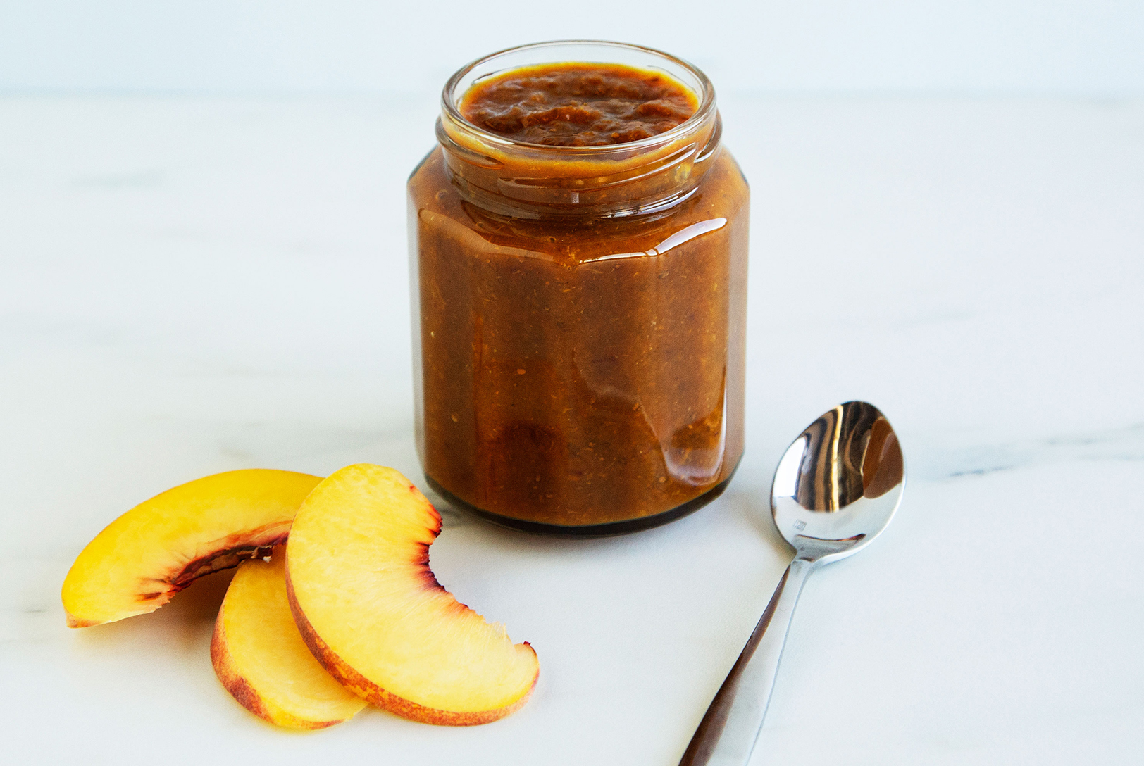 A glass jar filled with golden-brown Peaches and Cinnamon Chia Jam, next to two fresh peach slices and a silver spoon on a white marble surface.