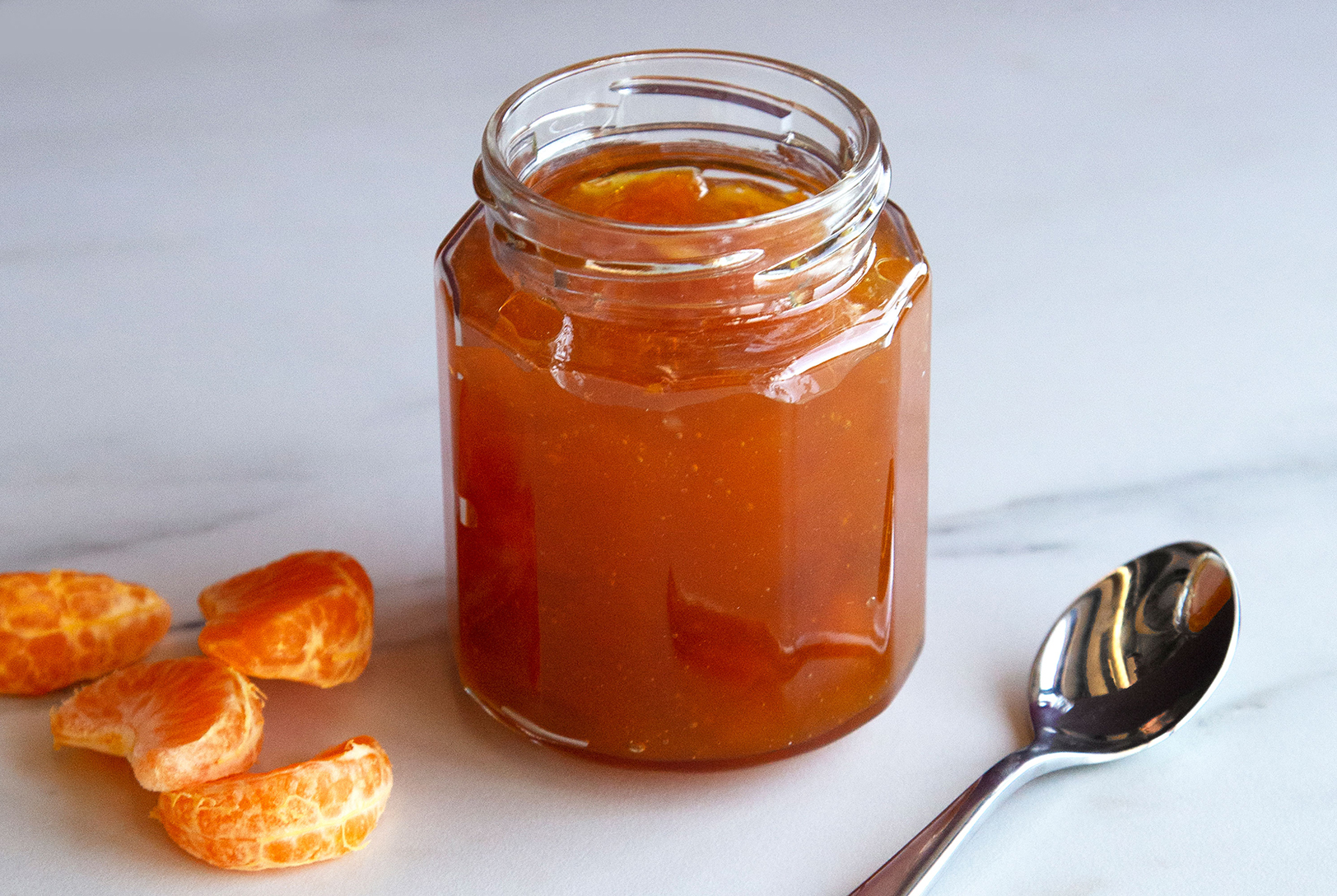 A glass jar filled with Caramelized Clementine Marmalade, with a spoon and clementine segments on a white marble surface.