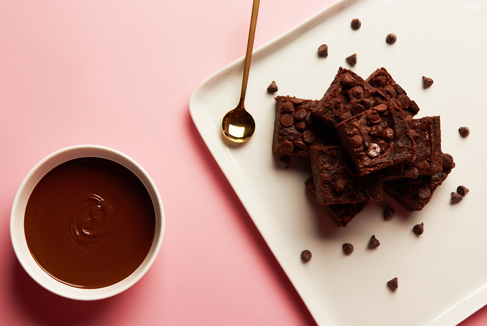 Black Bean Brownies topped with chocolate chips on a white plate, next to a bowl of rich chocolate sauce.