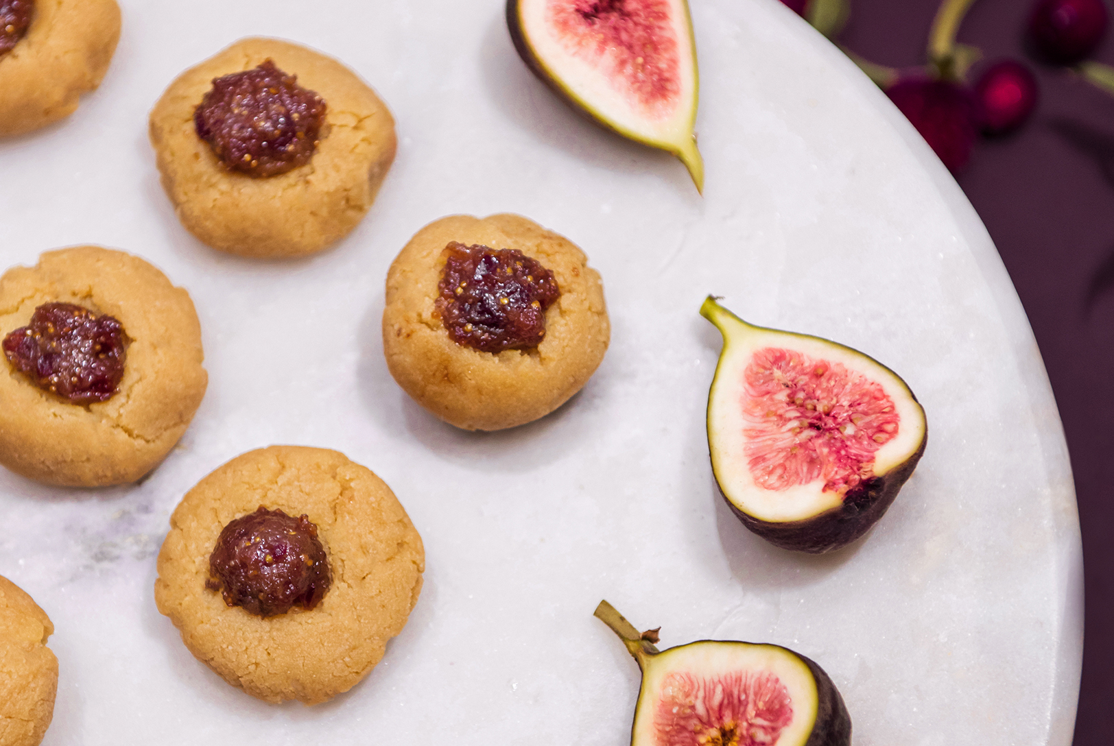 Several golden almond thumbprint cookies topped with dark cranberry fig jam on a white marble surface, alongside sliced fresh figs.