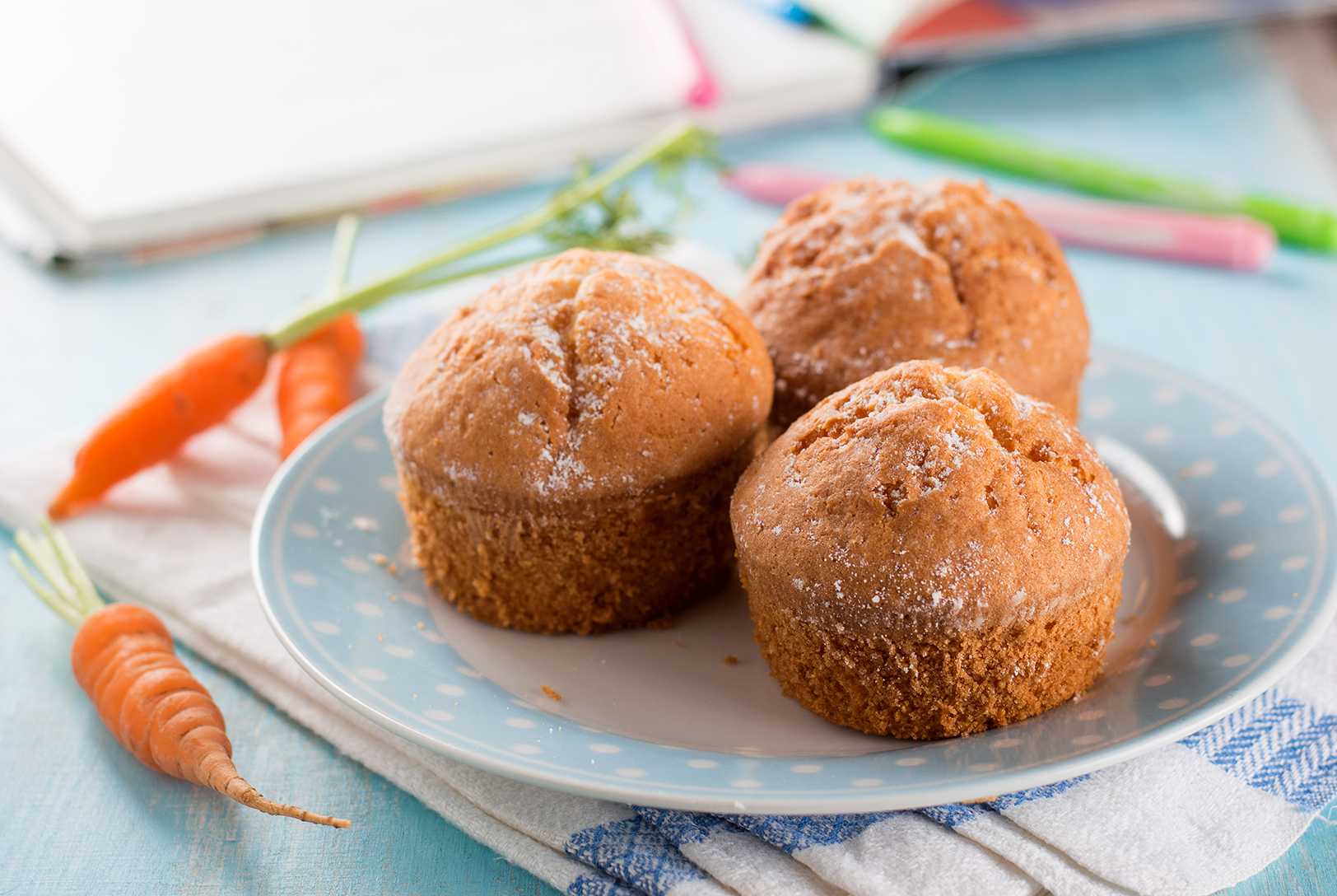 Three Apple Carrot Muffins dusted with sugar on a light blue polka dot plate, with fresh baby carrots nearby over a cloth napkin.