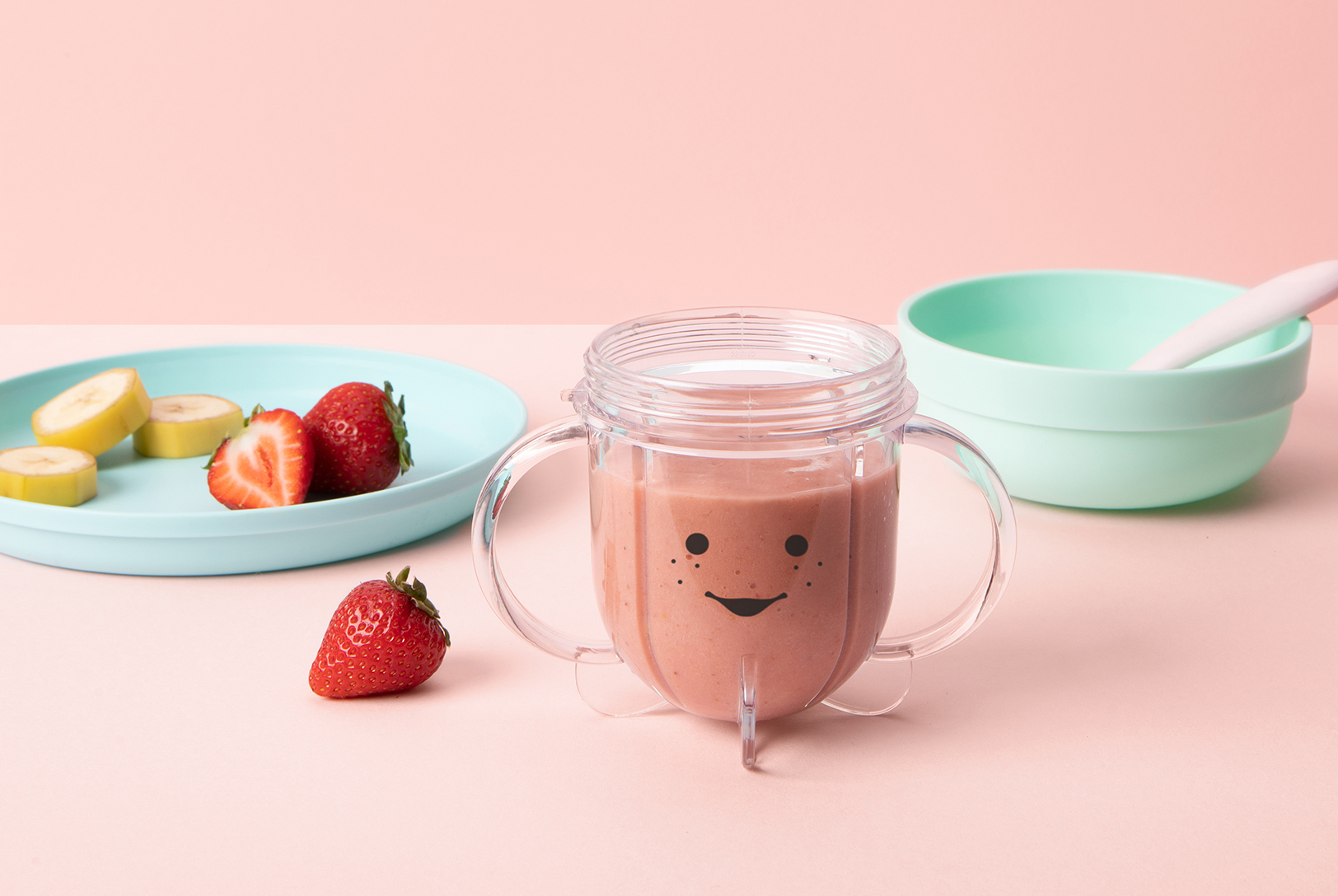 A small blender cup with a smiley face with Strawberry and Banana Purée, next to a plate with sliced bananas and strawberries, and a bowl with a spoon.