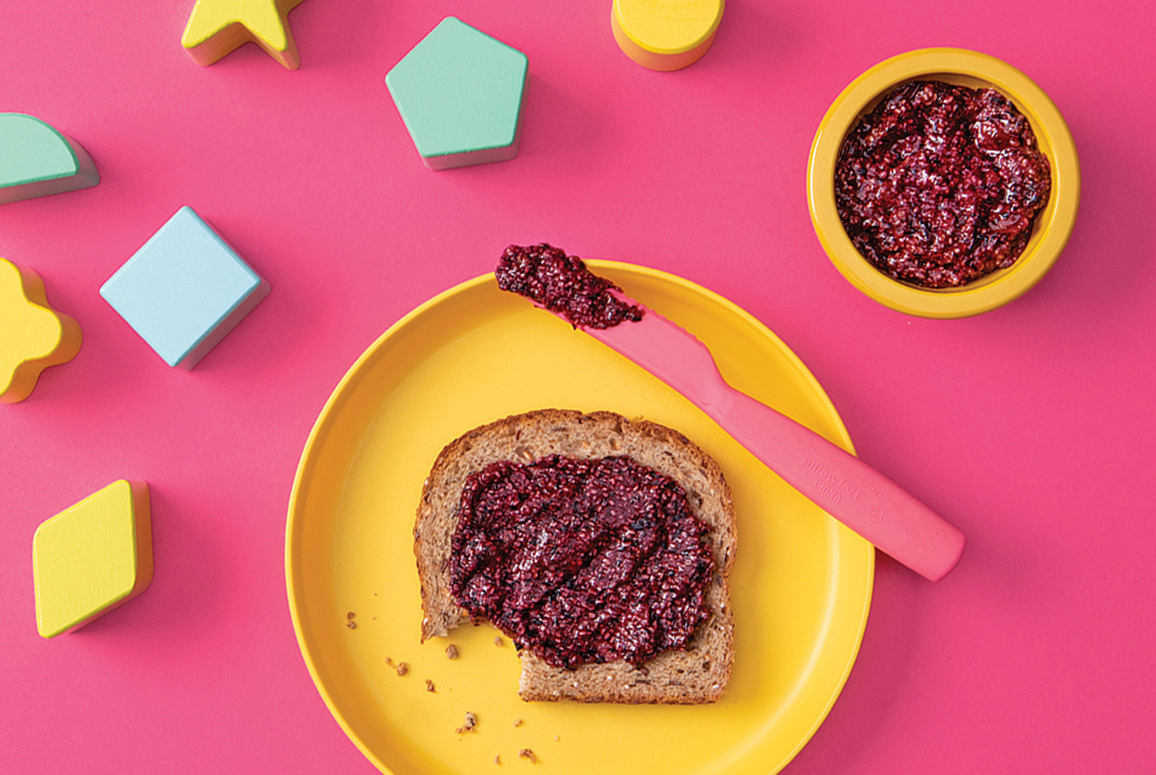 Overhead shot of Berry Chia Spread on a slice of toast with a pink knife, alongside a bowl of the spread and colorful wooden block toys.