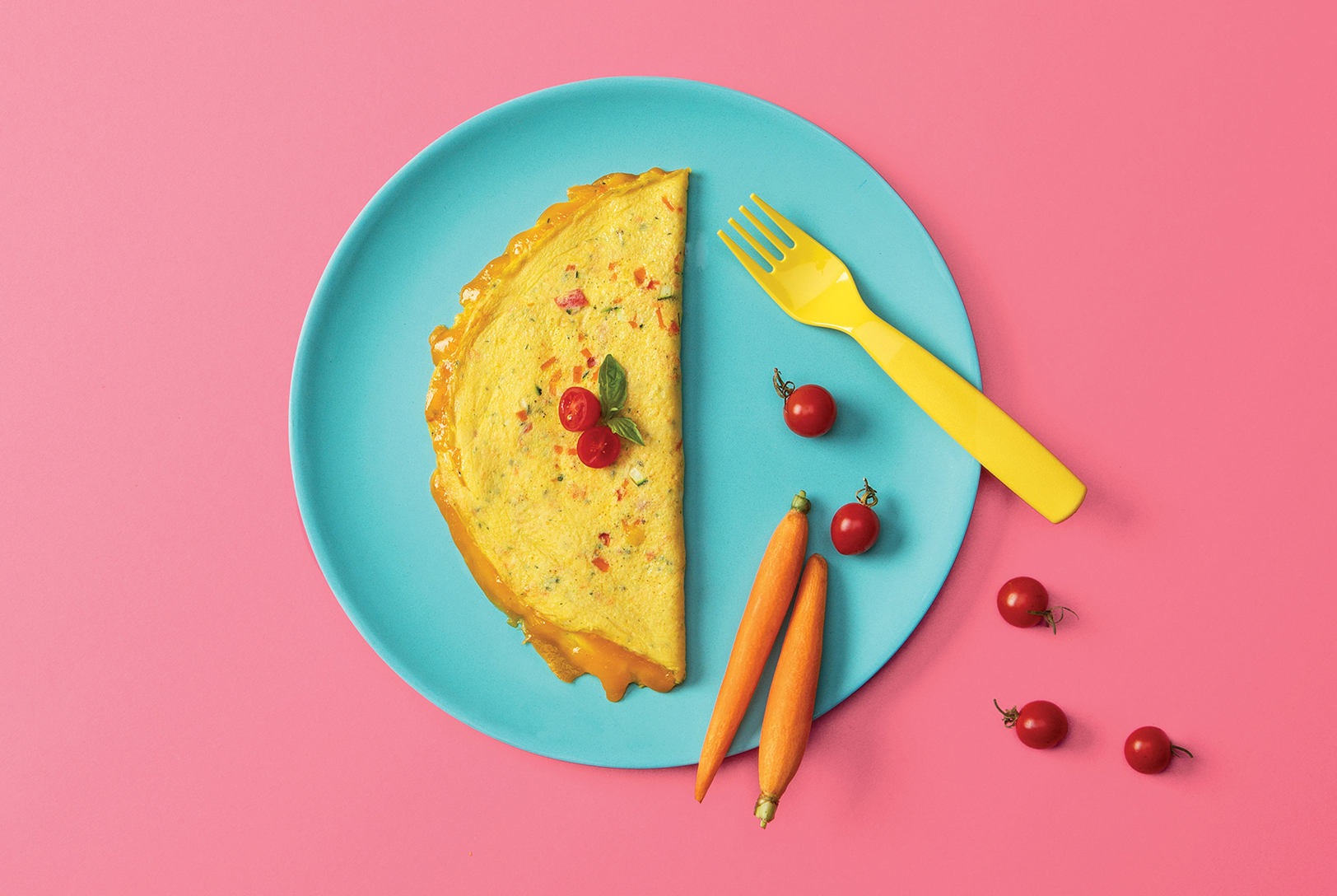 A Veggie Omelet on a bright blue plate, with cherry tomatoes, a yellow fork and small carrots on a pink background.