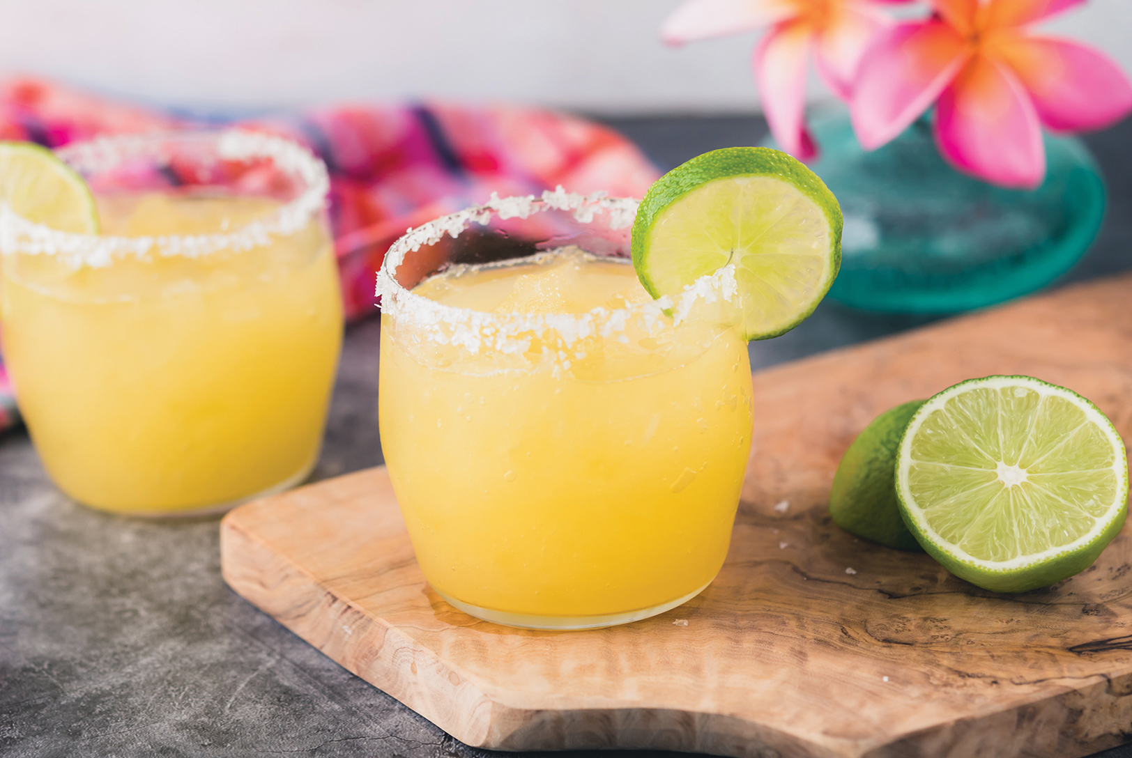 Two glasses of Frozen Margaritas with salted rims and lime wedges, on a wooden board with lime halves.