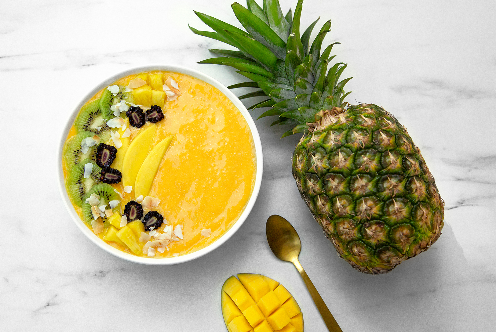Mango Pineapple Smoothie Bowl topped with fresh mango, kiwi slices, blackberries, and coconut flakes, next to a whole pineapple and a spoon.