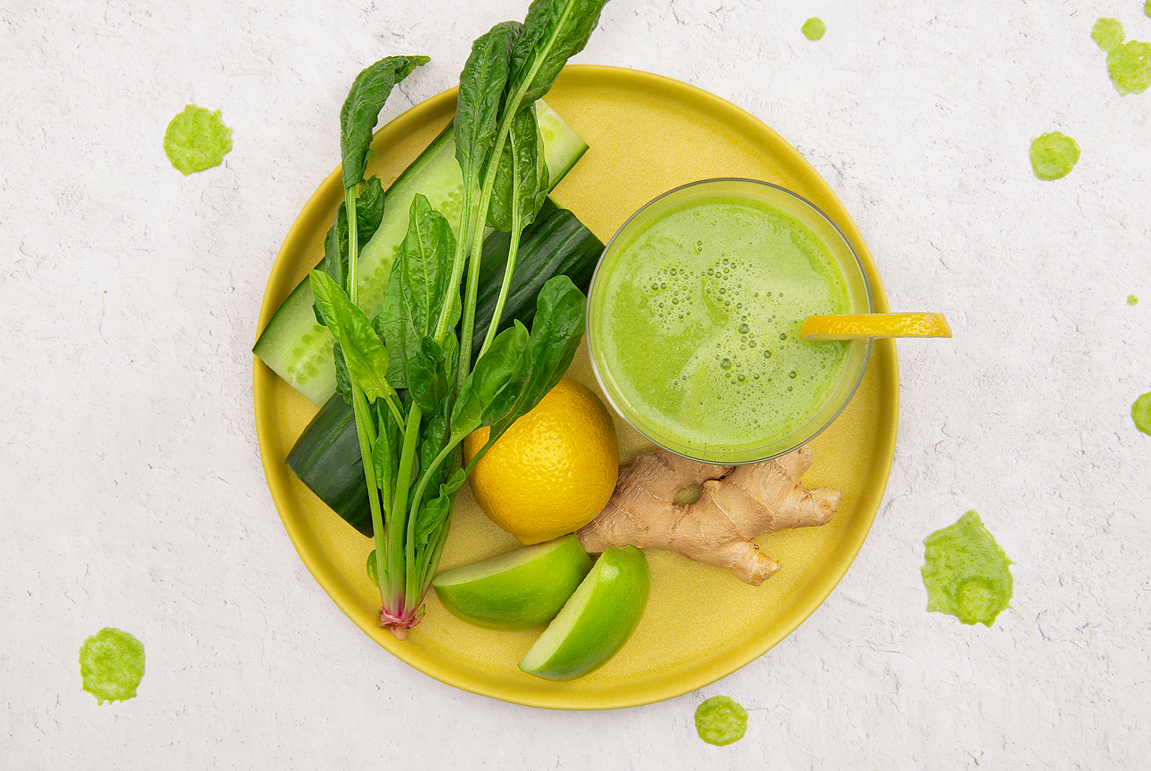 A green smoothie in a glass with a lemon wedge, surrounded by spinach, cucumber, a whole lemon, sliced green apples, and ginger on a yellow plate.