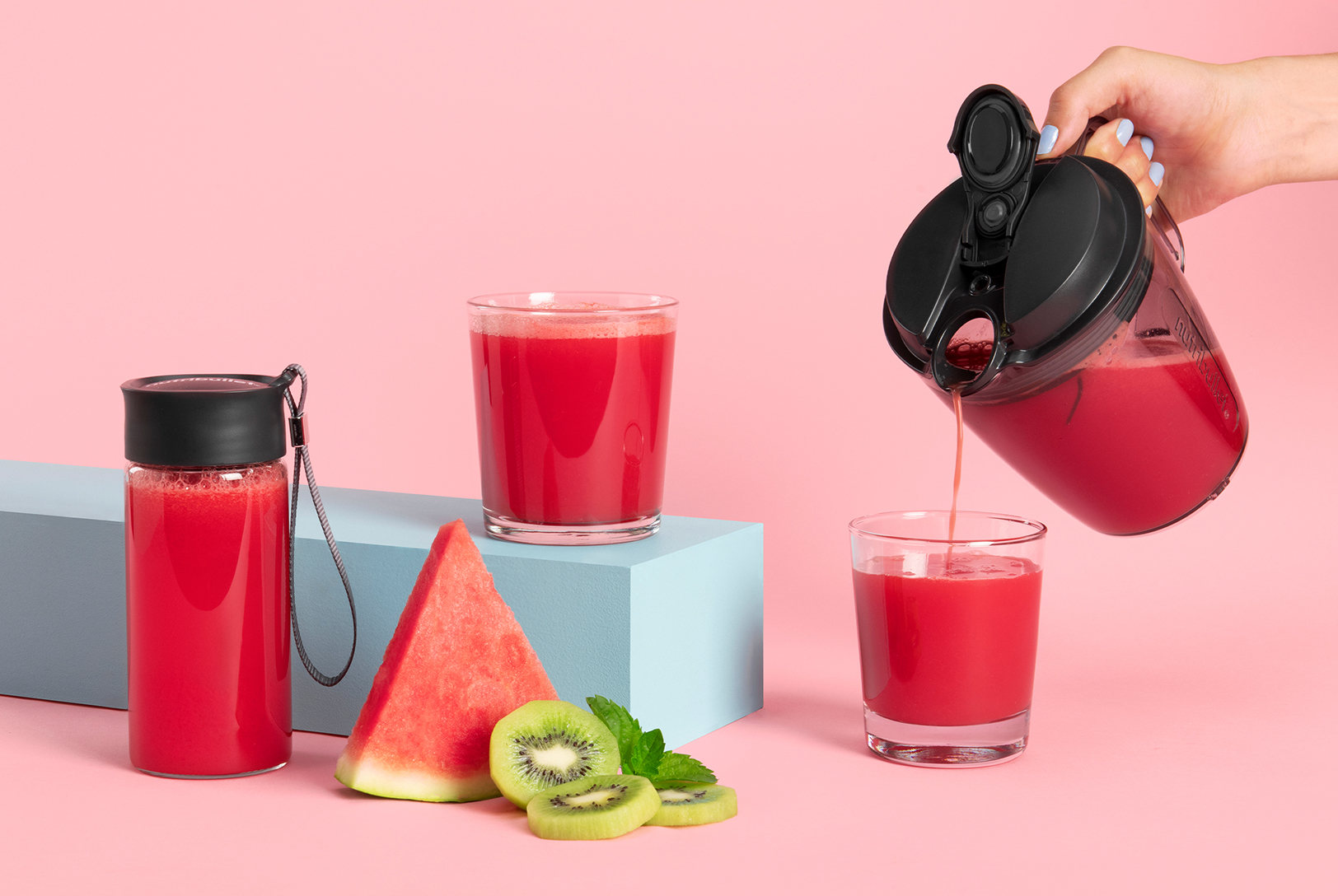 A hand pouring Watermelon Kiwi Nectar into a glass, with another filled glass and blender cup, along with watermelon and kiwi, on a pink background.