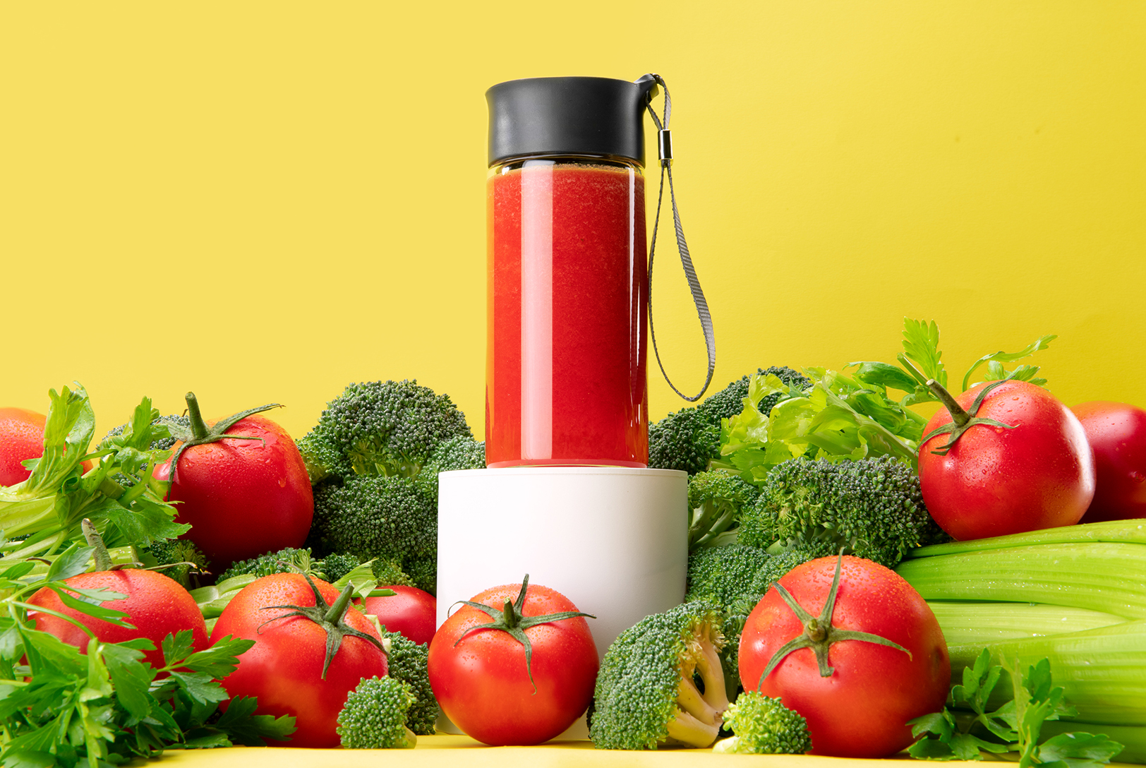 A bottle of red vegetable juice surrounded by tomatoes, broccoli florets, celery stalks, and lettuce leaves on a bright yellow background