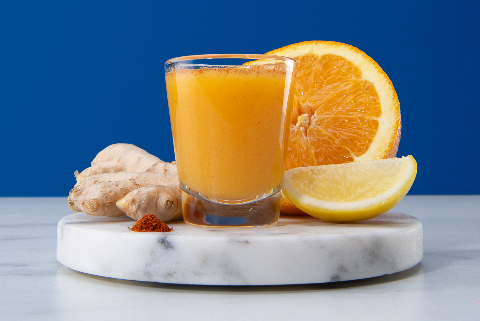 Ginger Zinger Shot in a glass with fresh ginger, orange, and lemon on a round marble coaster against a deep blue backdrop.
