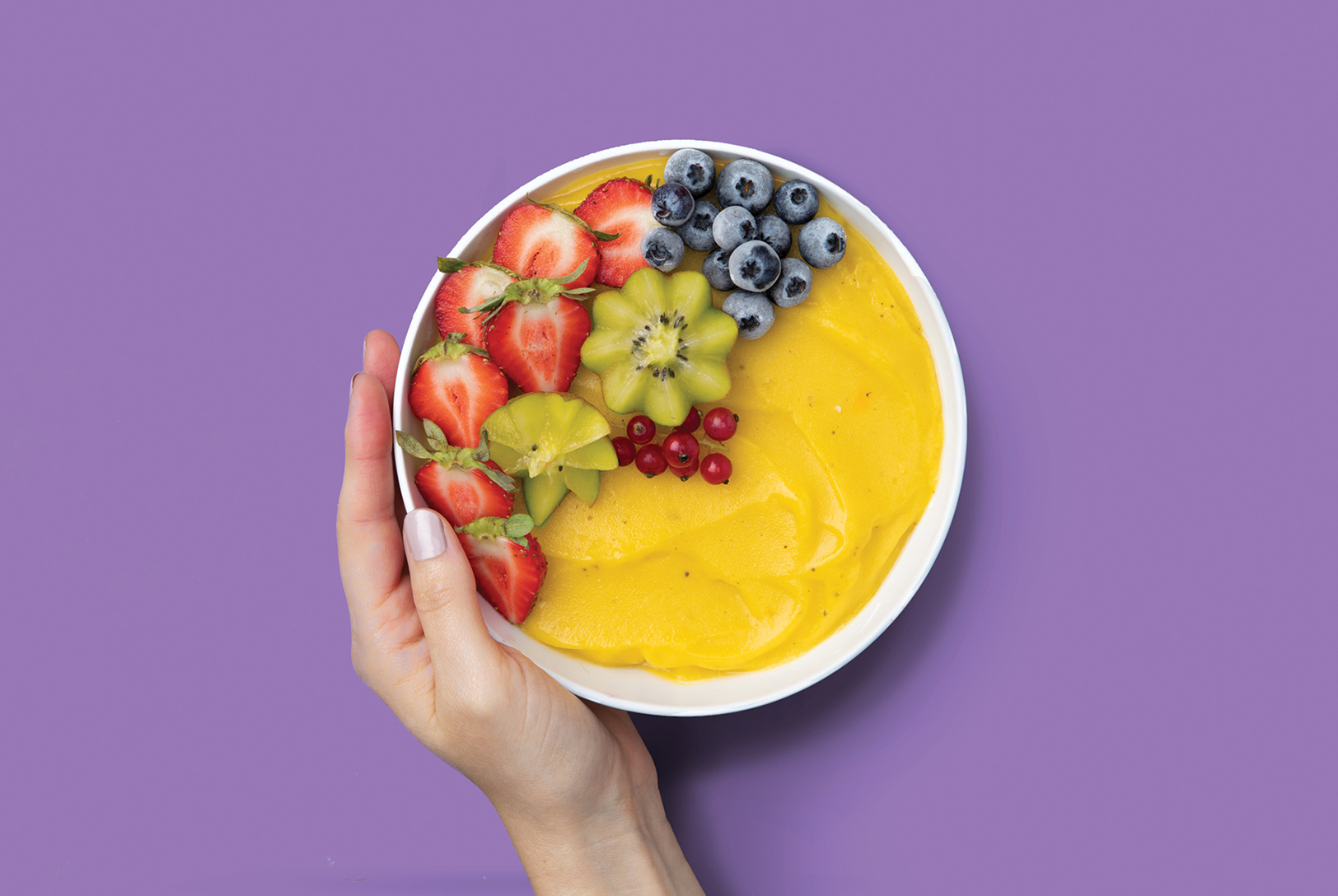 hand holding bowl of yellow stone fruit smoothie with fresh berries and kiwi