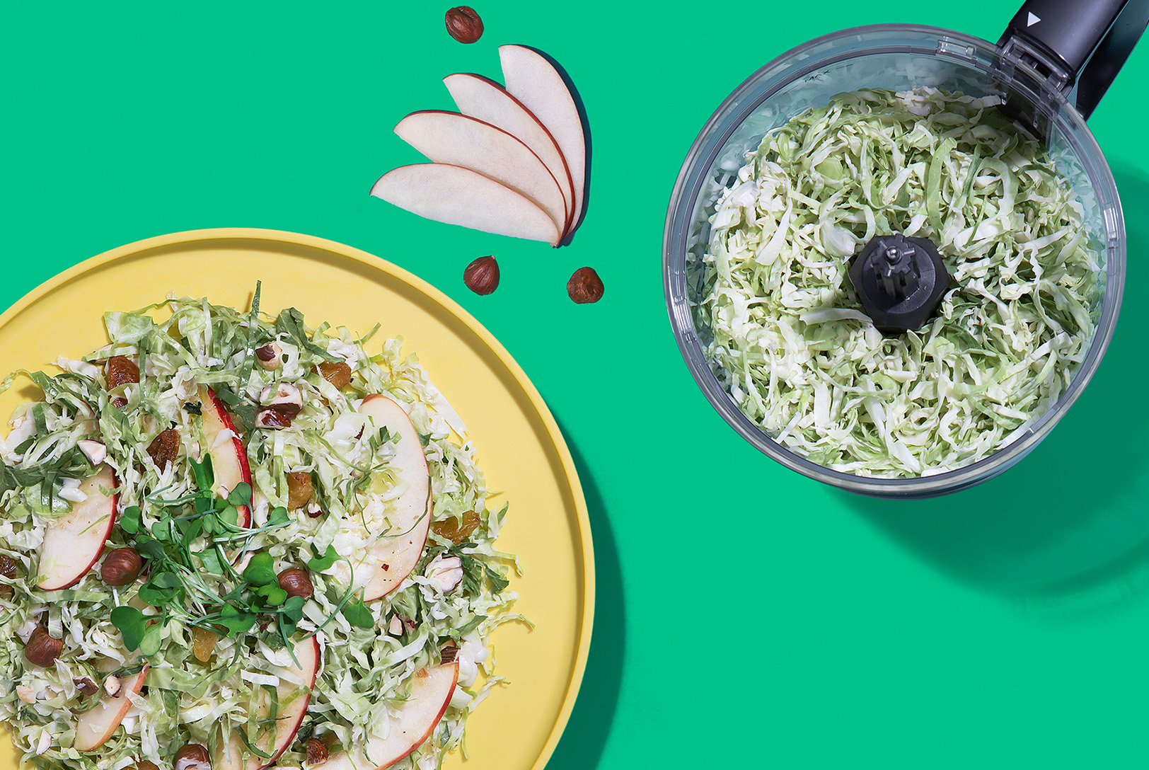 plate of shaved brussels sprouts salad with apples, nuts and raisin next to food processor