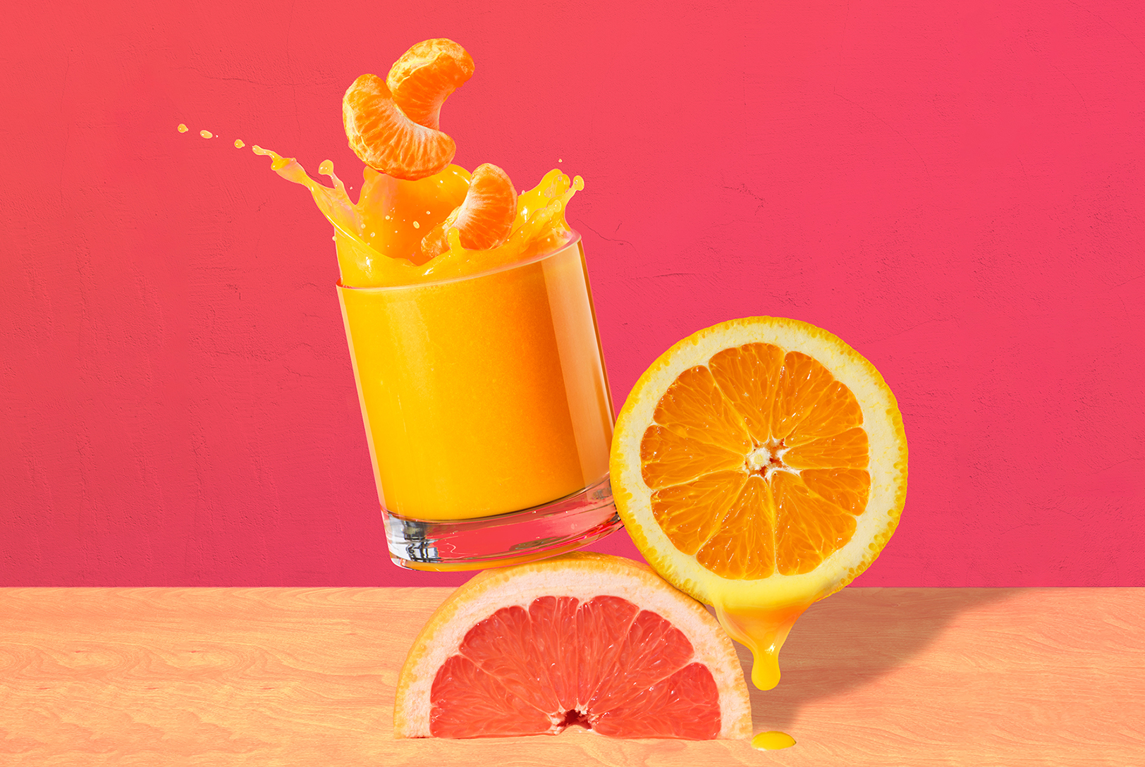 A glass of orange, clementine and grapefruit juice sitting on top of grapefruit with clementine splashing in glass in front of salmon colored background