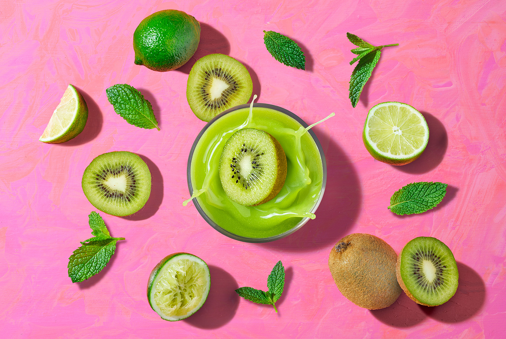 glass of kiwi, mint, lime juice with fruits around in front of a pink background