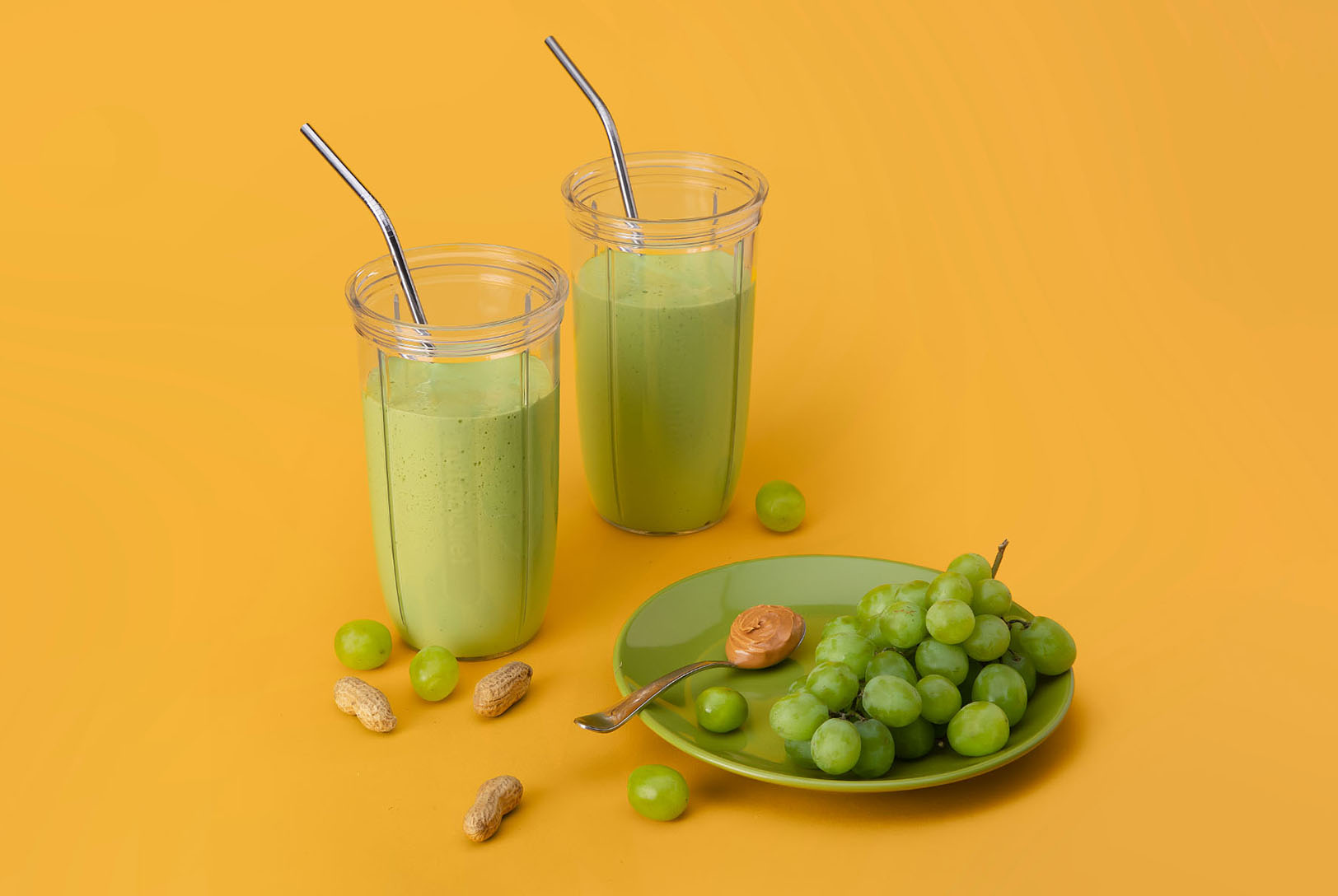 green smoothie in nutribullet cups on yellow background surrounded with fresh grapes, peanuts, and a spoonful of peanut butter
