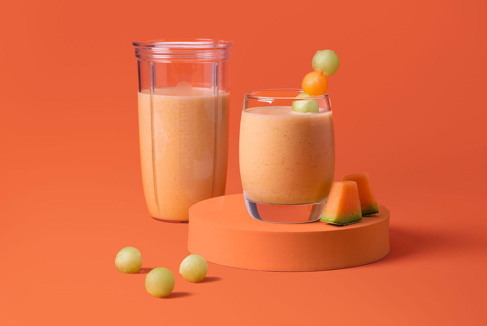 Orange melon cocktail in nutribullet cup and glass garnished with melon ball skewer on orange background