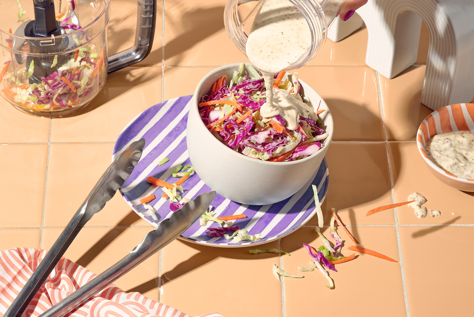 A bowl of colorful shredded red cabbage and carrots with creamy dressing being poured over it, with a food processor and tongs nearby.
