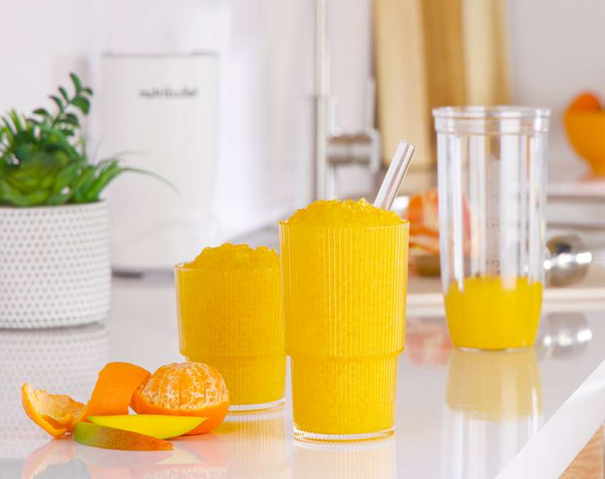Two orange mango slushies sit on a white counter beside fruit peels, a blender cup, and a nutribullet Turbo, creating a fresh and vibrant kitchen scene.