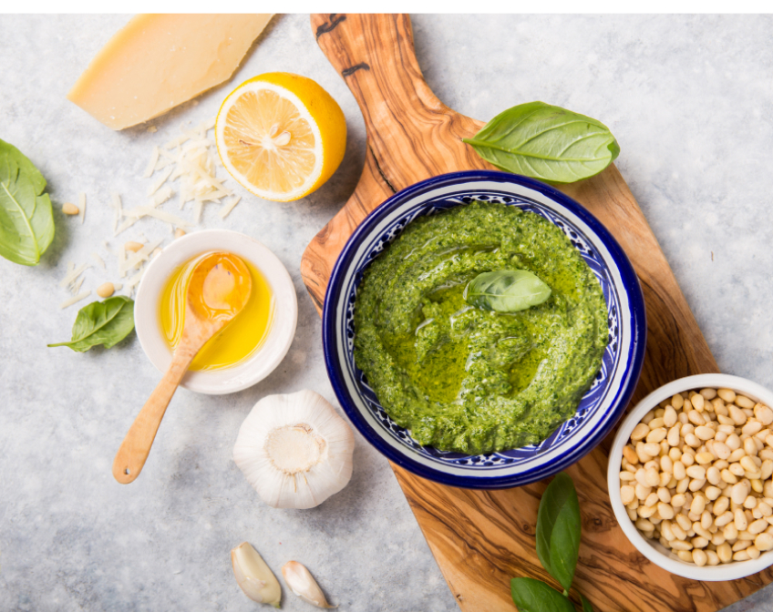 Image of a pesto lying on a wodden cutting board surrounded by a variaety of food.