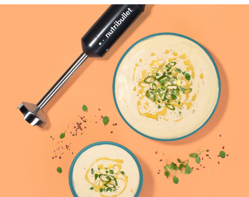 Image of the nutribullet Lite Immersion Blender lying on a peach colored countertop, next to two bowls of food preparations.
