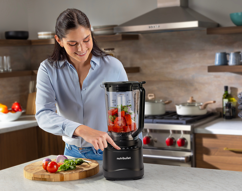Woman pressing power button on black nutribullet Power Core Blender with fresh tomatoes and vegetables inside.